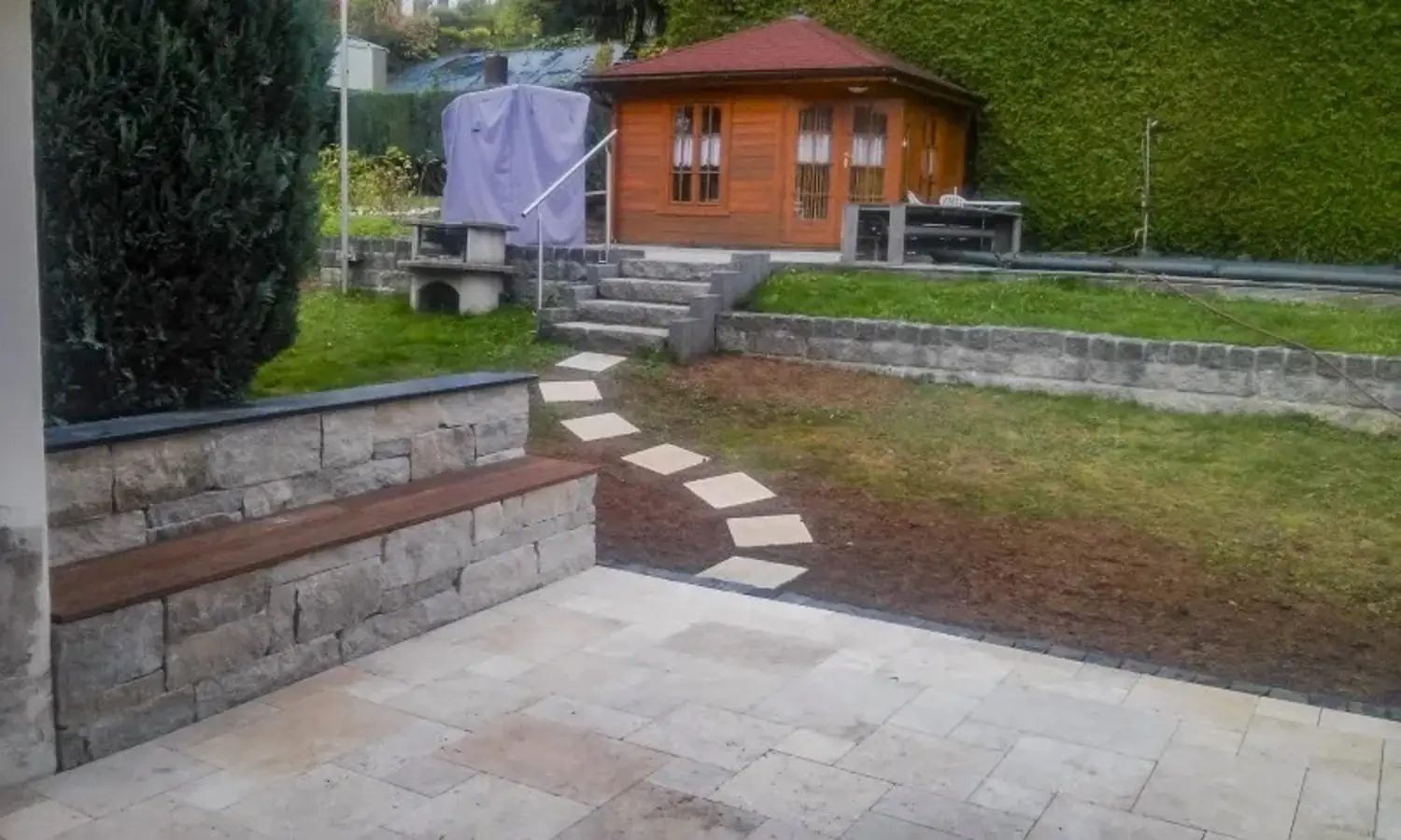 Garten mit gepflasterter Terrasse, Steinmauerbank und Gartenhaus auf einer Rasenfläche