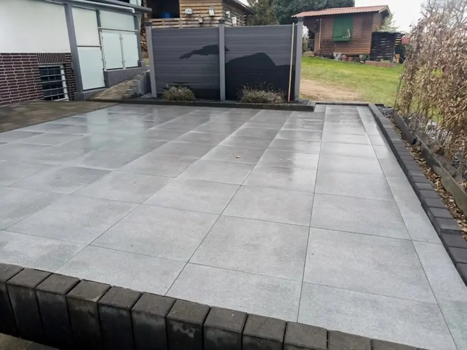 Gepflasterte graue Terrasse vor einem Haus mit Holzzaun