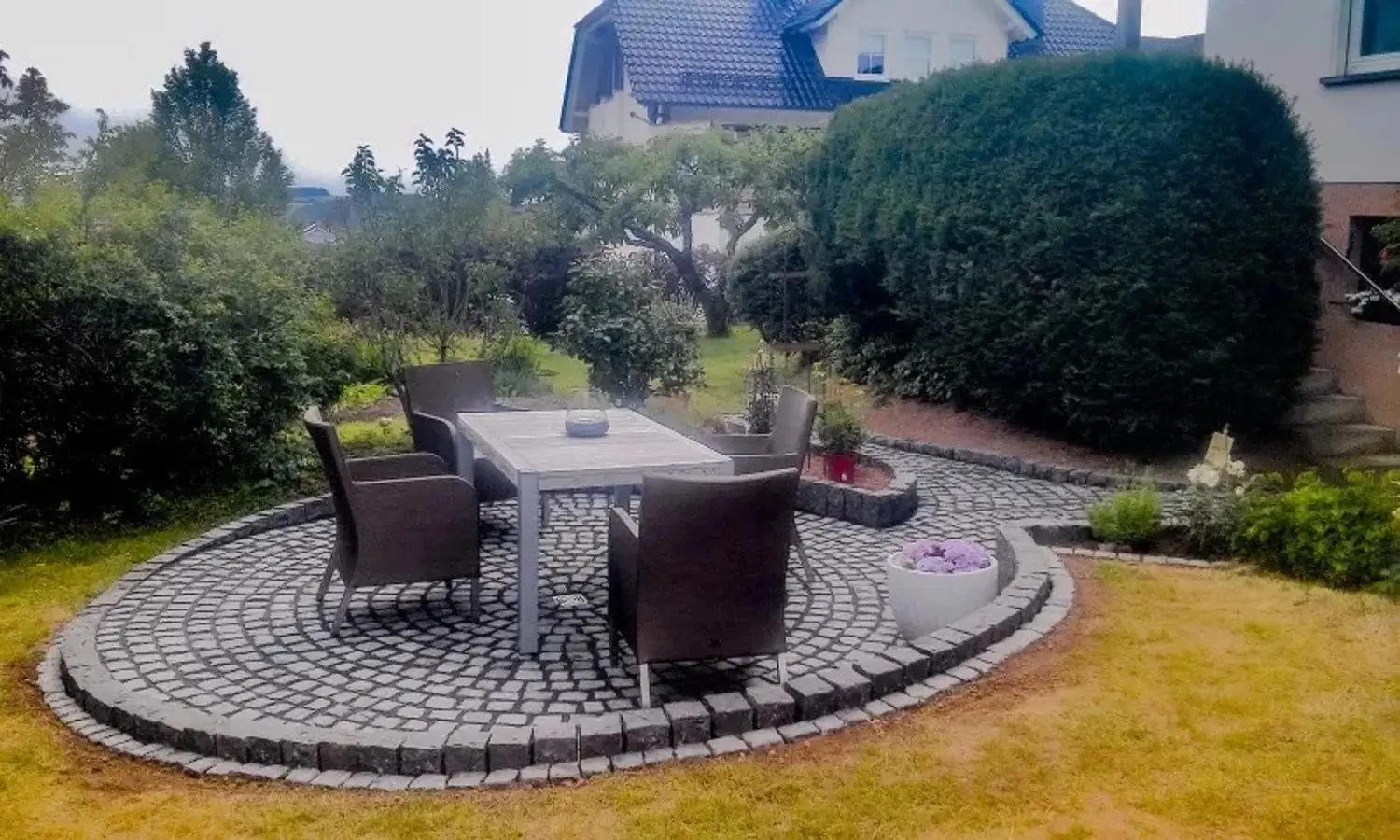 Ovaler Terrassenbereich aus Kopfsteinpflaster mit weißem Tisch und Stühlen